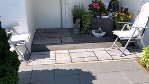 Um jardim frontal com um pátio pavimentado, uma porta branca, um pequeno jardim com pedras e plantas, e duas cadeiras.