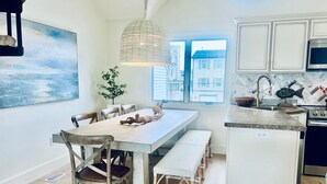 Une cuisine moderne avec des meubles blancs, un coin repas avec une table et des chaises en bois, un luminaire suspendu et une vue sur les bâtiments par la fenêtre.