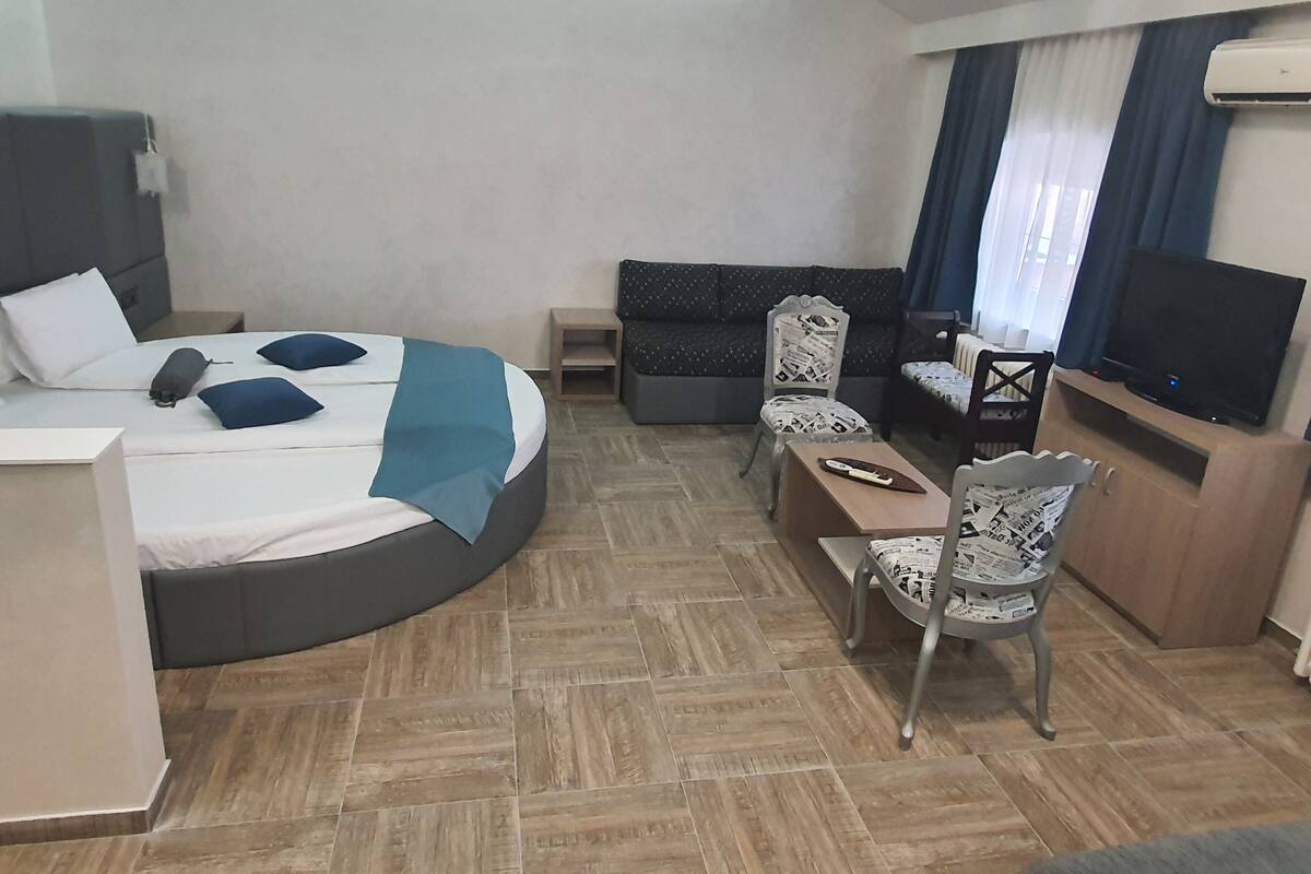 A modern hotel room with a curved bed, a sofa, a TV, and a dining table.