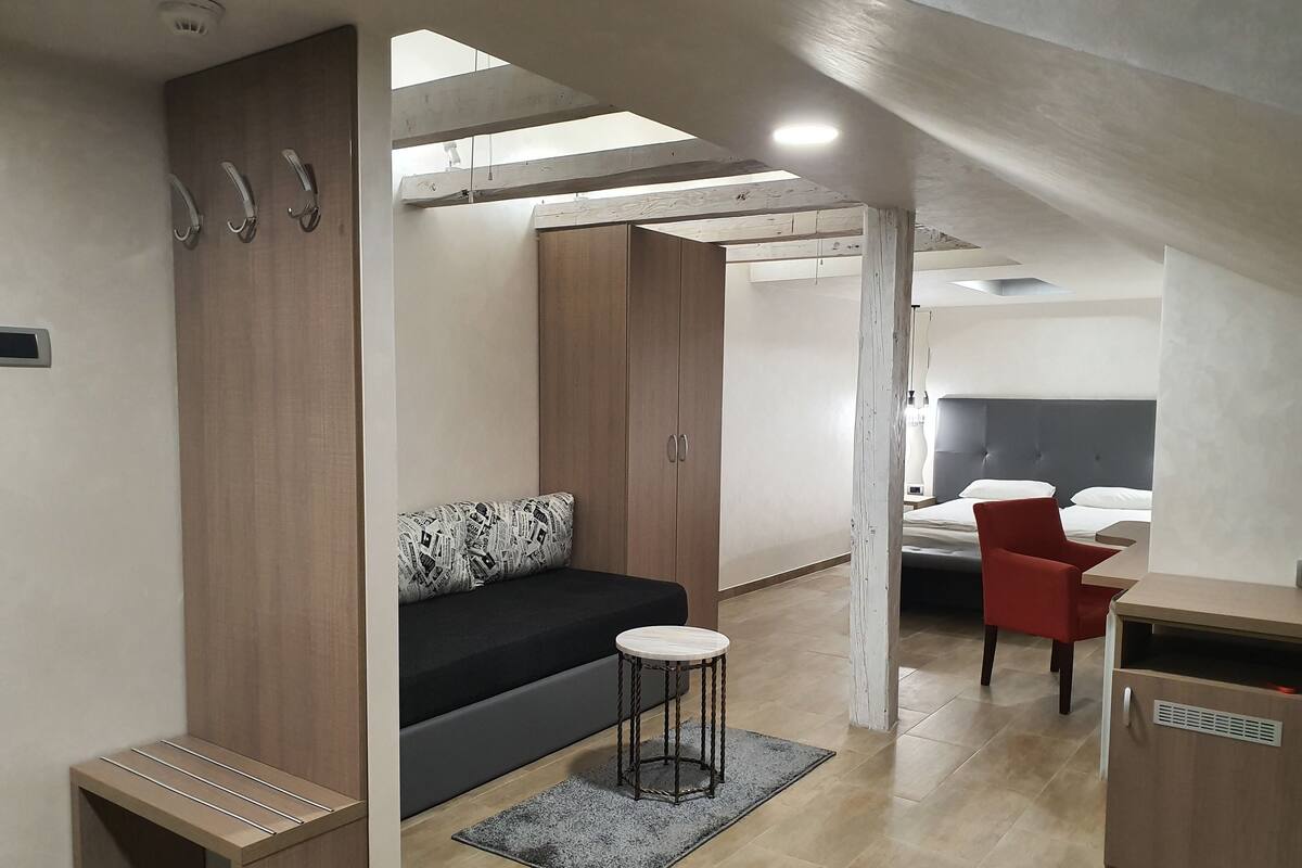 A modern, compact room with a sofa, a small table, and a bed with a red chair.