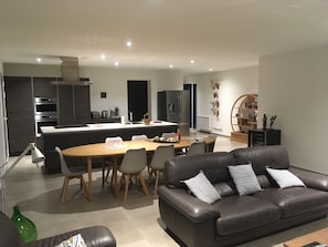 Un salon moderne avec un canapé en cuir, une table à manger et des chaises. On aperçoit au fond une cuisine équipée d’appareils électroménagers et dotée d’un petit îlot central.