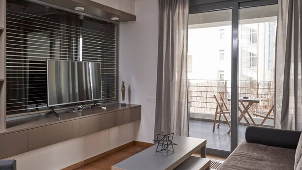 En moderne stue med et fladskærms-tv, et sofabord og en sofa. Der er en balkon med udendørs møbler, som kan ses gennem vinduet.