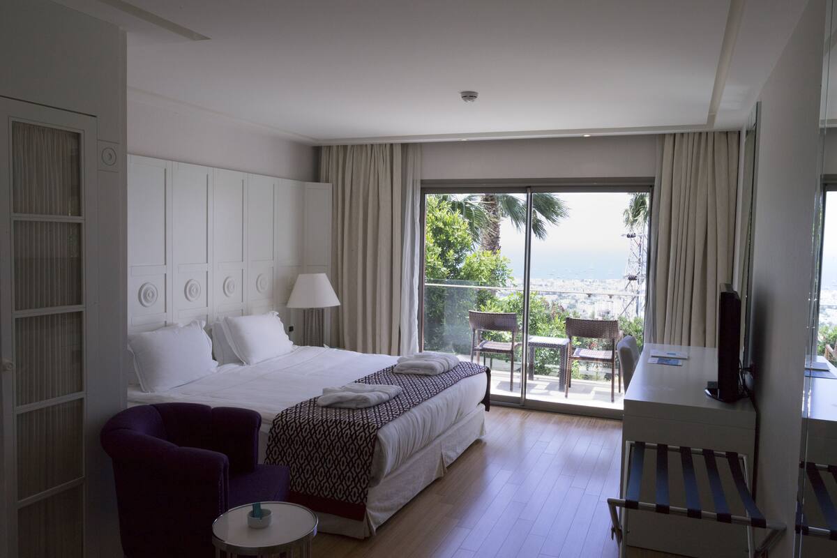 A hotel room with a large bed, a desk, a chair, and a balcony with a view.