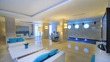 Un moderno vestíbulo de hotel con un mostrador de recepción, una zona de estar con sofás blancos y cojines azules, un cuadro grande en la pared y una piscina con fuente de agua.