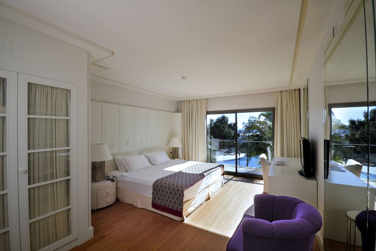 A modern hotel room with a large bed, a desk, a purple chair, and a balcony with a view of palm trees.