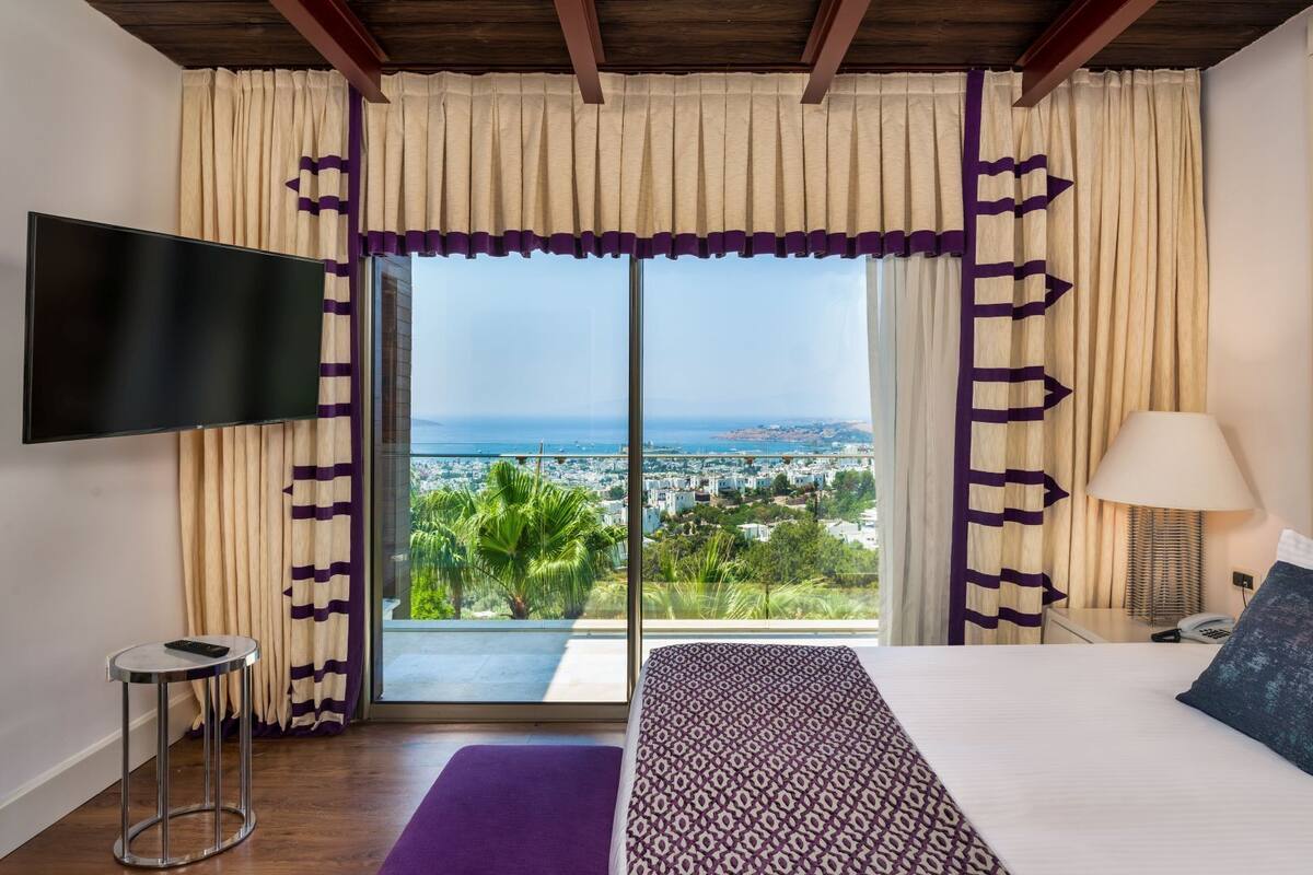 A hotel room with a bed, a TV, a nightstand, a lamp, and a view of a cityscape through a large window.