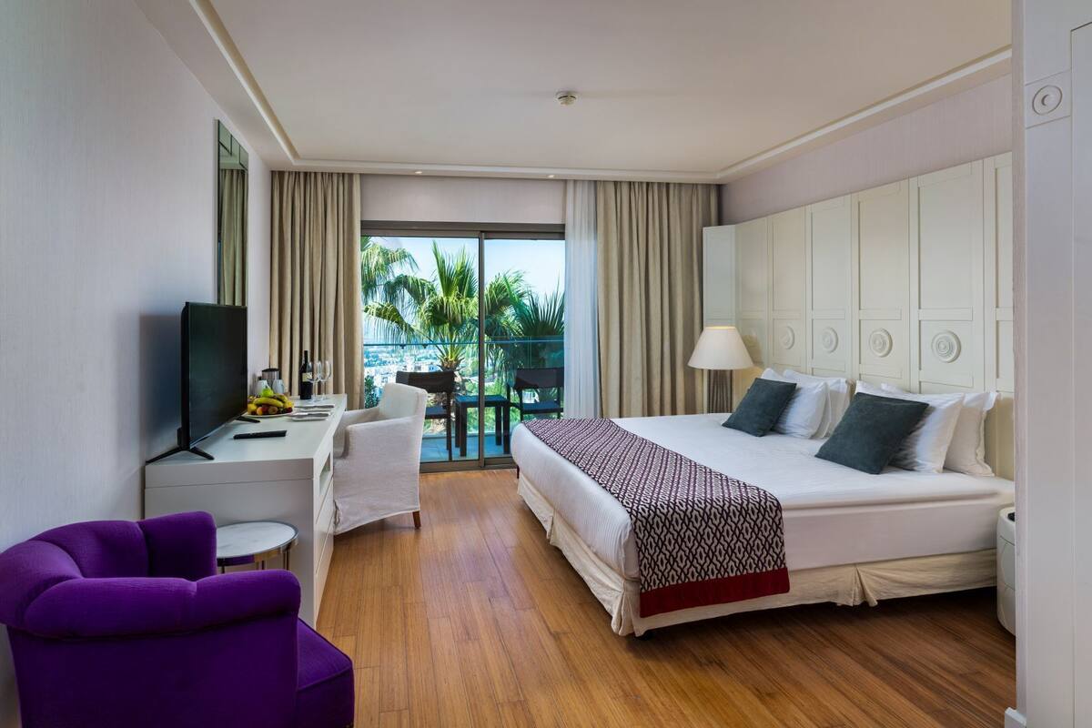 A hotel room with a bed, a desk, a chair, a TV, and a balcony with a view of palm trees.