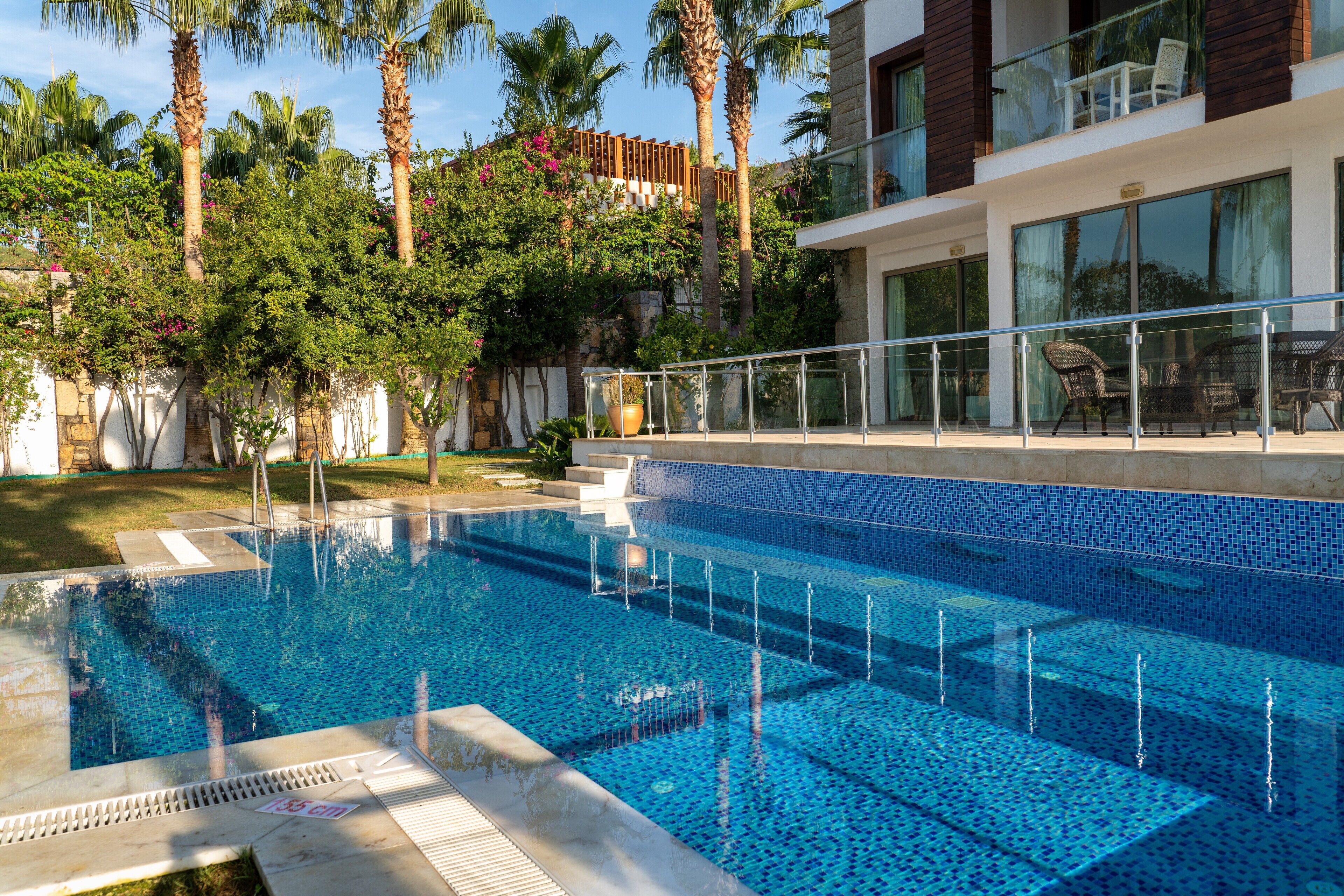 A swimming pool with clear blue water, surrounded by palm trees and a modern building with large windows and a balcony.