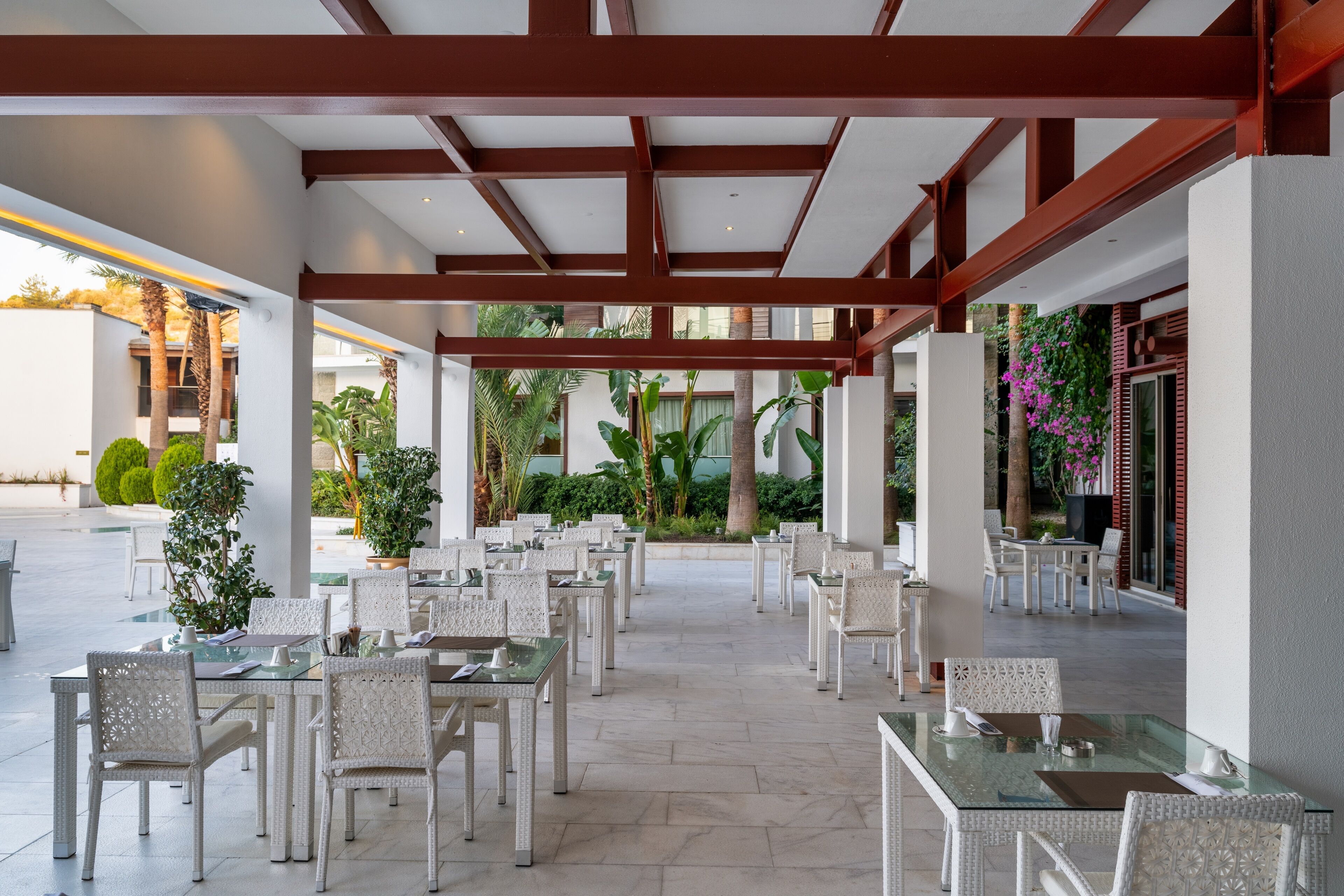 Un espace de restauration en plein air avec des tables en verre et des chaises en osier, entouré de verdure et bordé d’une allée couverte.