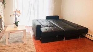 Un dormitorio con cama negra, paredes blancas, una mesita con una planta y una ventana con cortinas blancas.