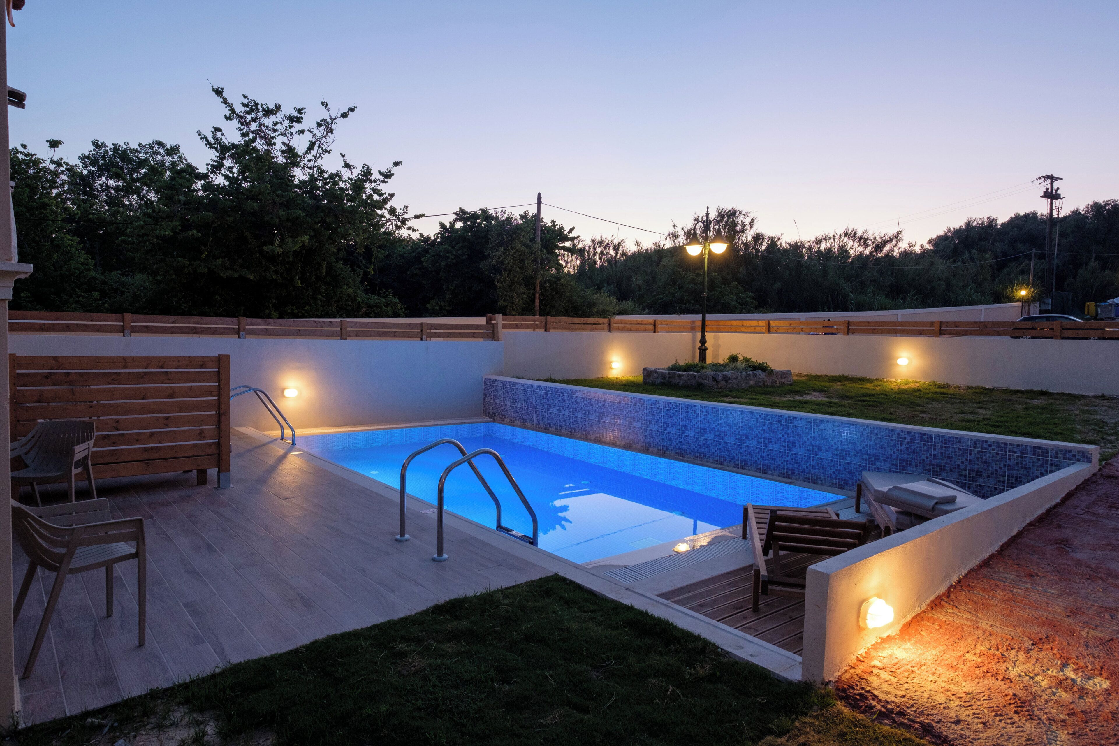 Una piscina rectangular al aire libre con borde de azulejos, rodeada por una terraza de madera con sillas y una pequeña mesa.
