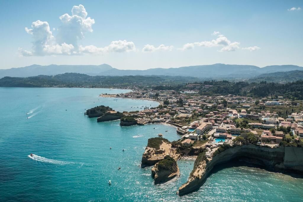 An aerial view of a coastal town with a beach, clear blue sea, and distant mountains.