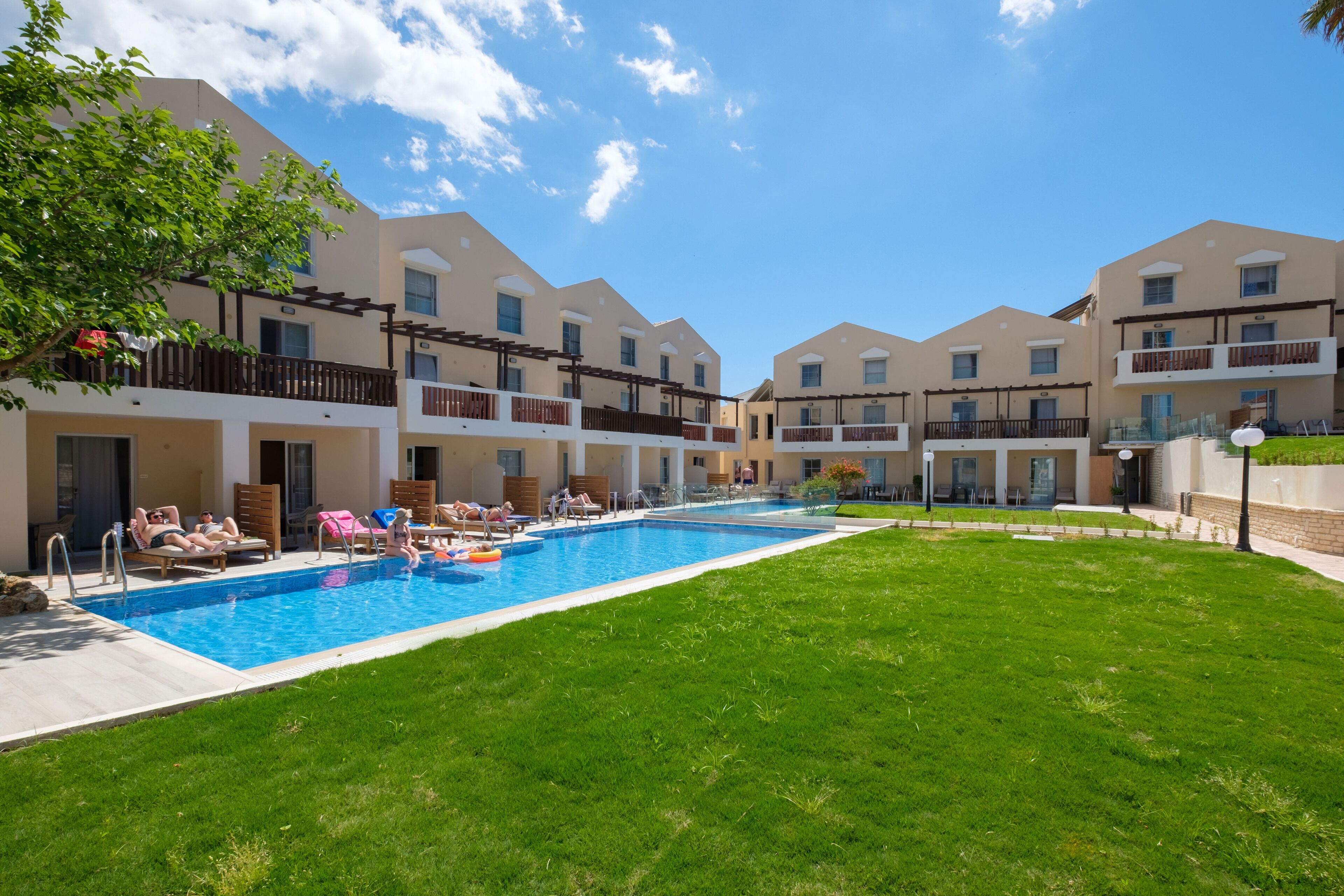 A swimming pool surrounded by a well-maintained lawn, with a building featuring balconies and a clear blue sky.