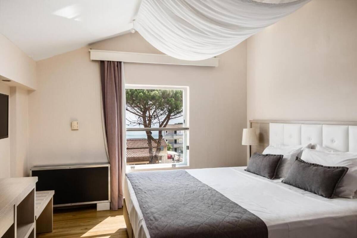 Een slaapkamer met een bed, een raam met gordijnen, een radiator en uitzicht op gebouwen.