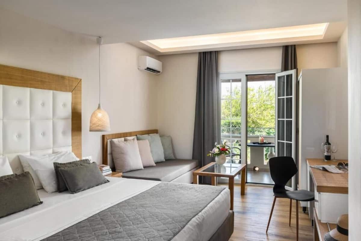 Een moderne hotelkamer met een groot bed, een bank, een kleine tafel en een stoel. Er is een raam met gordijnen en uitzicht op groen.