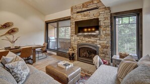 A cozy living room with a stone fireplace, a TV mounted above it, and large windows offering a view of the outdoors.