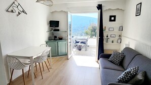A modern living room with a grey sofa, a white dining table with chairs, a kitchen area with a TV, and a view of the outdoors. - STUDOTEL 405 LES GENETS (Mollans-sur-Ouvèze)