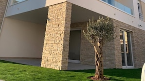 Un edificio moderno de dos plantas con base de piedra, amplios balcones de vidrio y un pequeño árbol en primer plano.