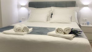 Una cama bien tendida con sábanas blancas y dos toallas enrolladas, un cabecero azul oscuro y dos luces fijadas a la pared.