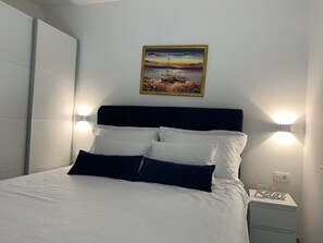Cama bem arrumada com roupa de cama branca e azul escura, cabeceira escura e duas luzes de leitura fixadas na parede.