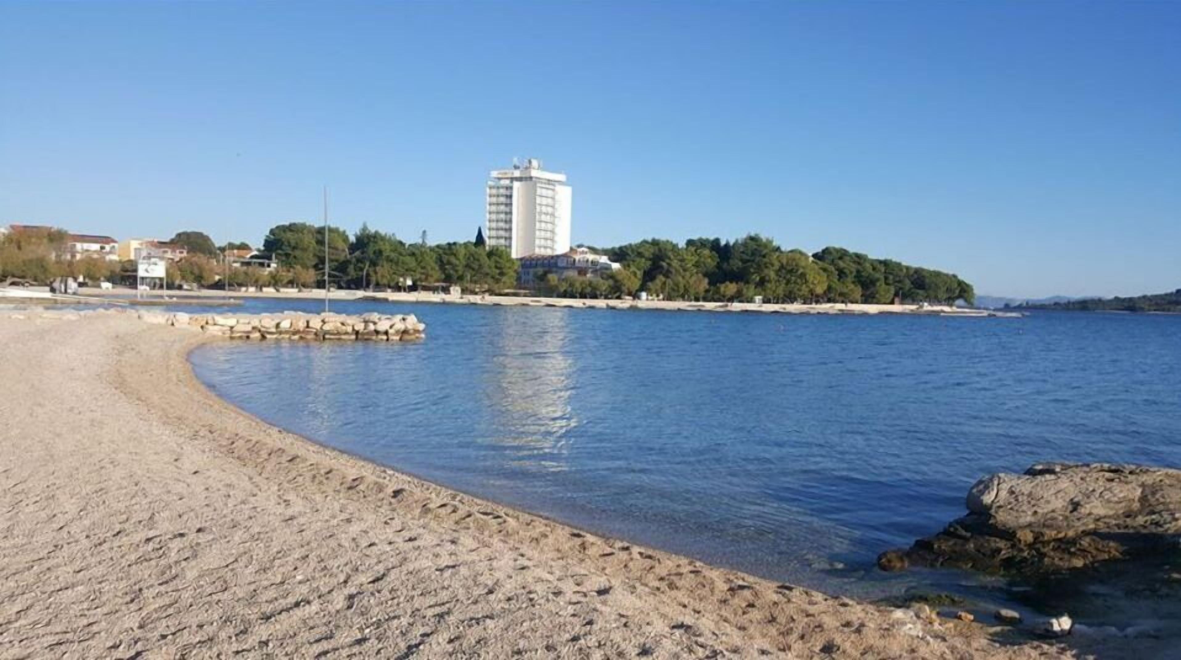 A sandy beach with calm water, a rocky outcrop, and a building in the background.