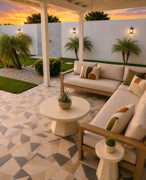 A covered outdoor seating area with a sofa and two round tables, surrounded by a geometric patterned floor and a white wall with a sunset view. - NEW! MODERN GLAM RESORT STYLE OLD TOWN SCOTTSDALE DUAL PRIMARY BDRMS HEATED POOL (Scottsdale)