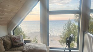 A cozy living room with a large window overlooking a beach, a wooden ceiling, and a telescope on a tripod.