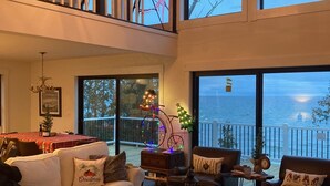 A spacious living room with a high ceiling, large windows offering a view of the sea, and a balcony with a railing.