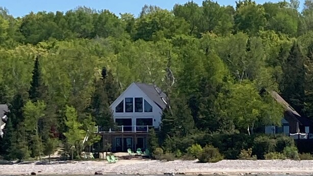 A two-story house with a large balcony, surrounded by dense forest and a body of water.