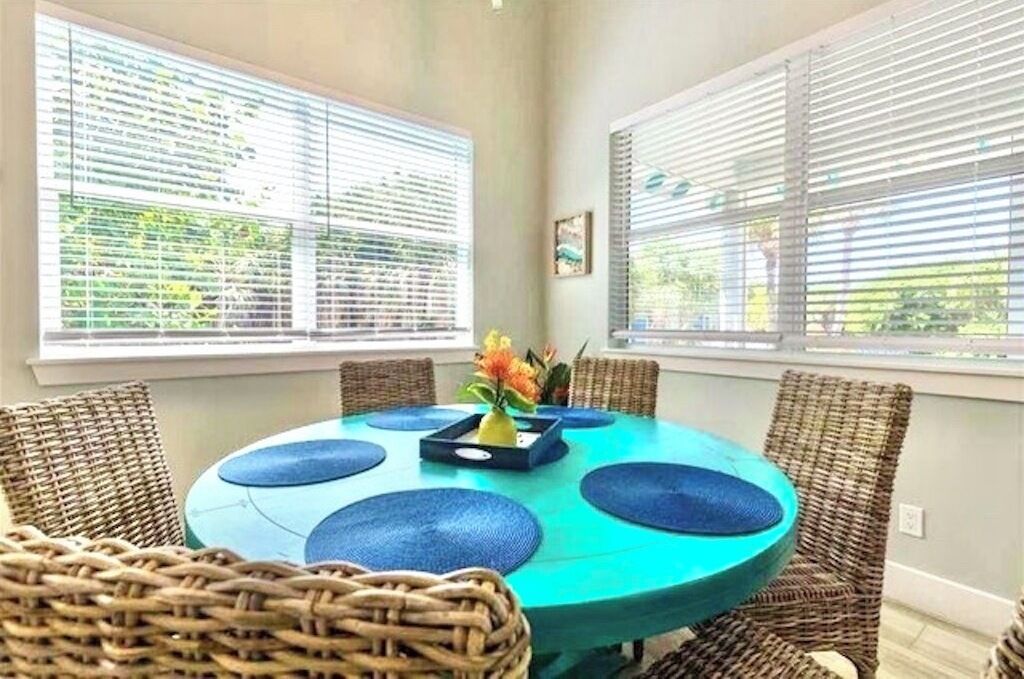 A dining area with a round table, blue placemats, wicker chairs, and a vase with flowers.