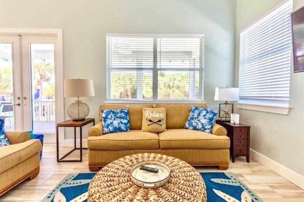 A living room with a beige sofa, two blue and white patterned pillows, a round woven coffee table, and a lamp on a side table.