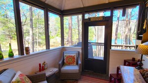Una habitación acogedora con una silla de mimbre, un banco acolchado y vistas al bosque a través de ventanas amplias. Se ve una puerta con un letrero que dice “Welcome to our Neck of the Woods”.