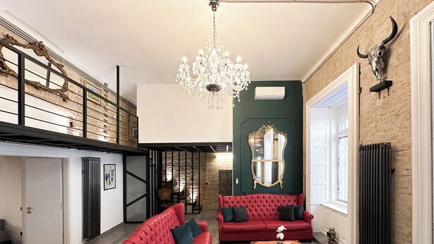 A spacious living area with a red sofa, a blue sofa, and a coffee table. A chandelier hangs from the ceiling, and a large mirror is mounted on the wall.