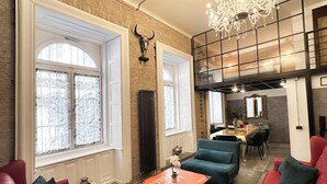 A spacious living area with a red sofa, teal armchairs, and a coffee table. A chandelier hangs from the ceiling, and a large window provides natural light.