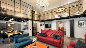 A spacious living area with a red sofa, blue armchairs, and a coffee table with a bold text design. A chandelier hangs from the ceiling, and a kitchen is visible in the background.