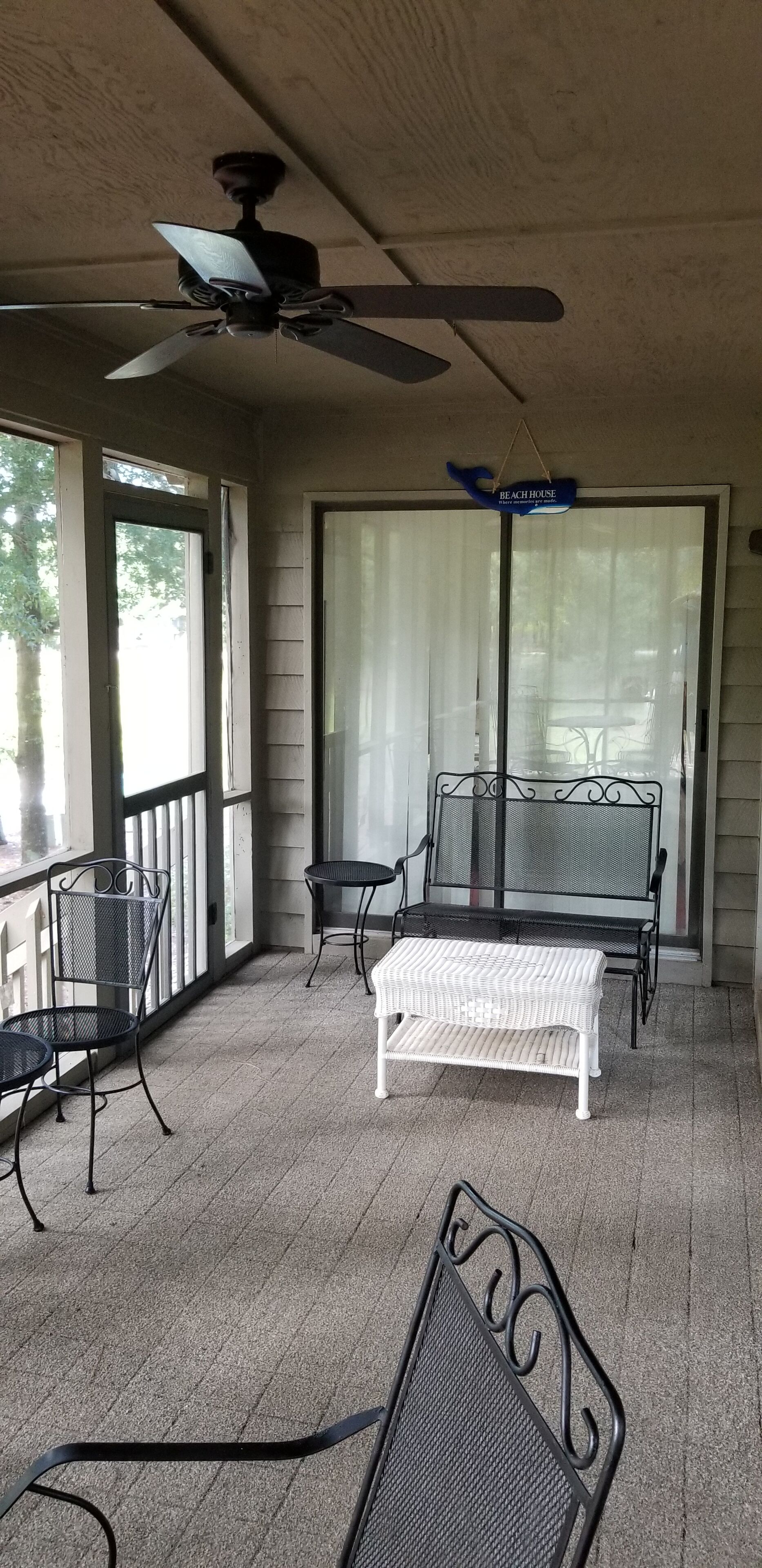 Un patio techado con ventilador de techo, dos sillas de metal, una mesa de mimbre blanco y una puerta de vidrio con un letrero que dice 'Beach House'.