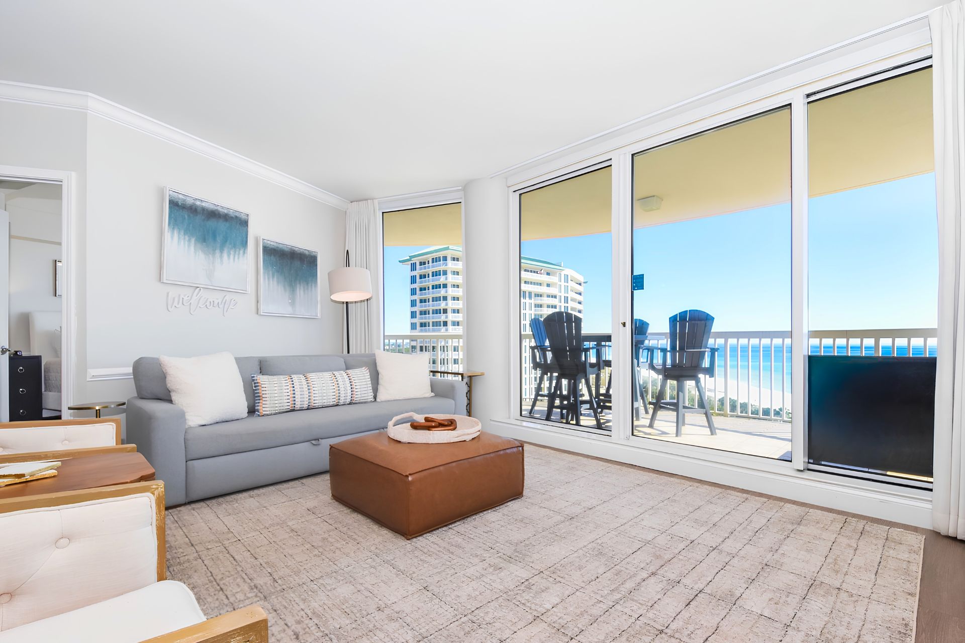 Ein modernes Wohnzimmer mit einem grauen Sofa, einem braunen Hocker und einem großen Fenster mit Blick auf ein Gebäude und das Meer.