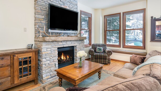 A living room with a stone fireplace, a television mounted above it, and a large window with a view of trees and a house. - Bluebird Day (Telluride)