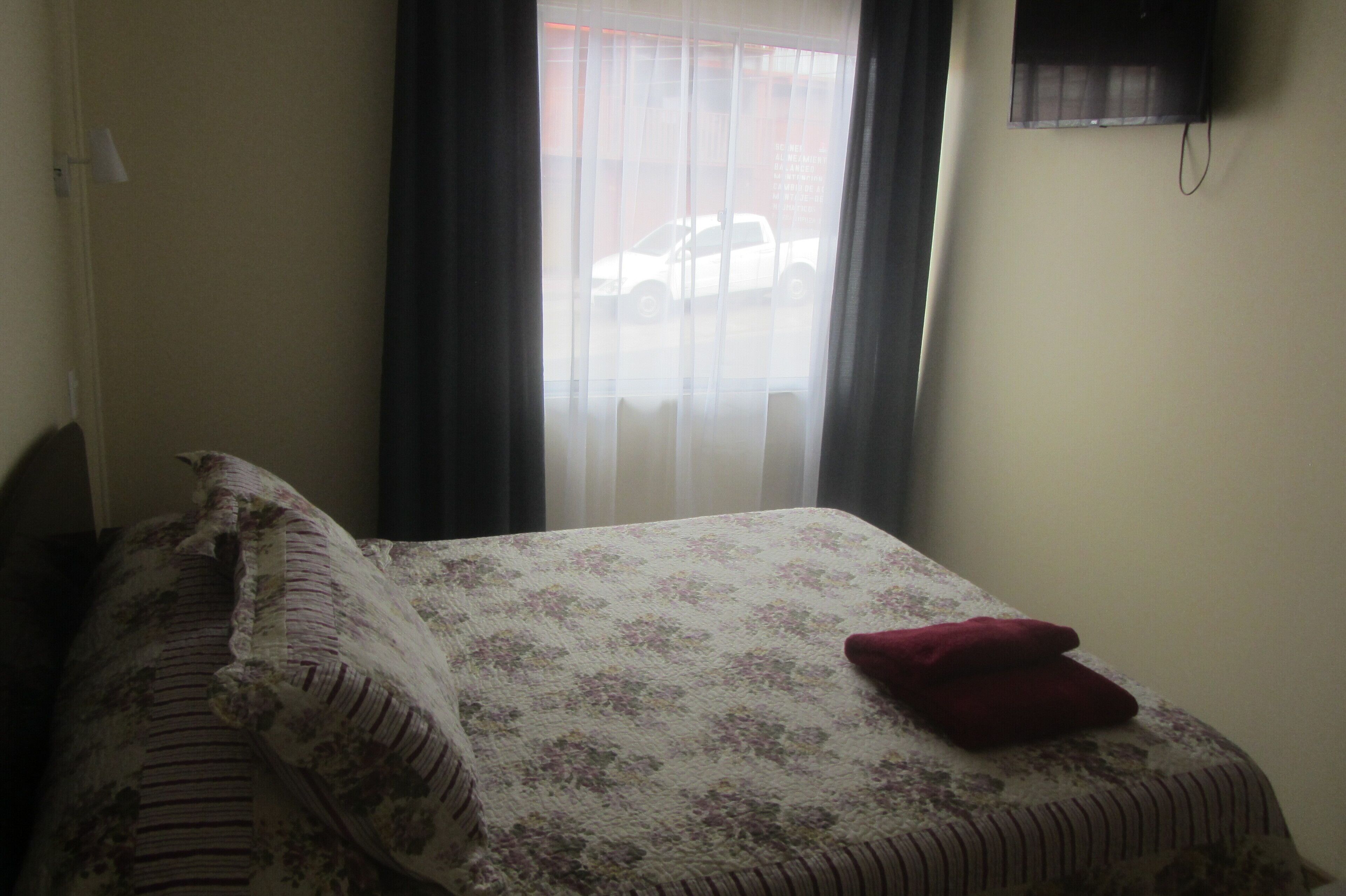 Un dormitorio con cama, ventana con cortinas y televisor montado en la pared.