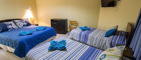Habitación de hotel con dos camas, cada una con ropa de cama y almohadas a rayas azul y blanco. Se observa una silla de madera y una mesita con una lámpara.