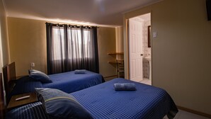 Hotellihuone, jossa on kaksi sänkyä, pieni pöytä, tuoli, ikkuna, jossa on verhot, ja ovi, joka johtaa kylpyhuoneeseen.