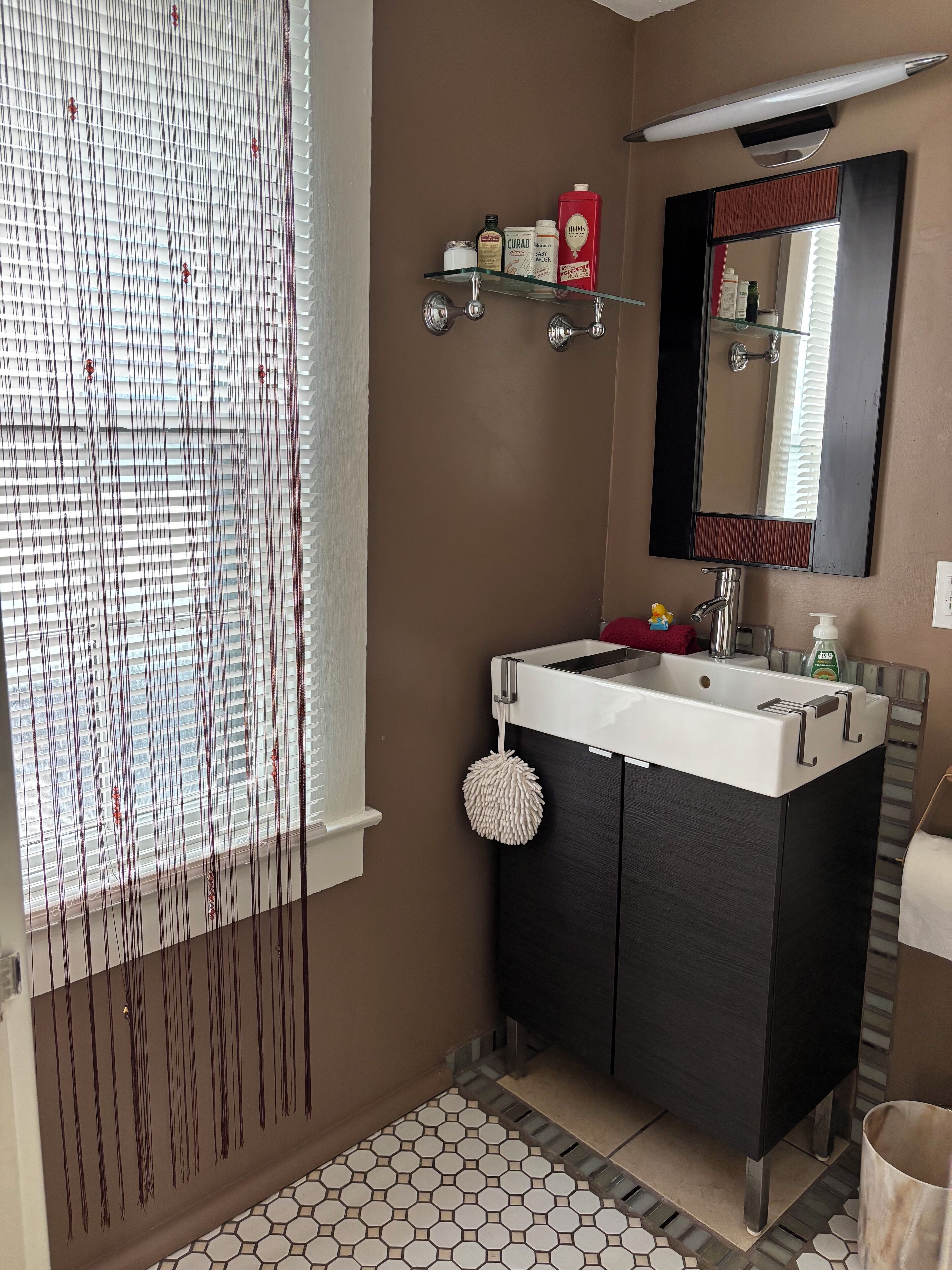 Une salle de bain avec un lavabo blanc, des meubles sombres, un miroir et une fenêtre avec des stores.