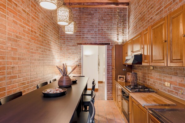 A kitchen with brick walls, wooden cabinets, a long dining table, and pendant lighting.