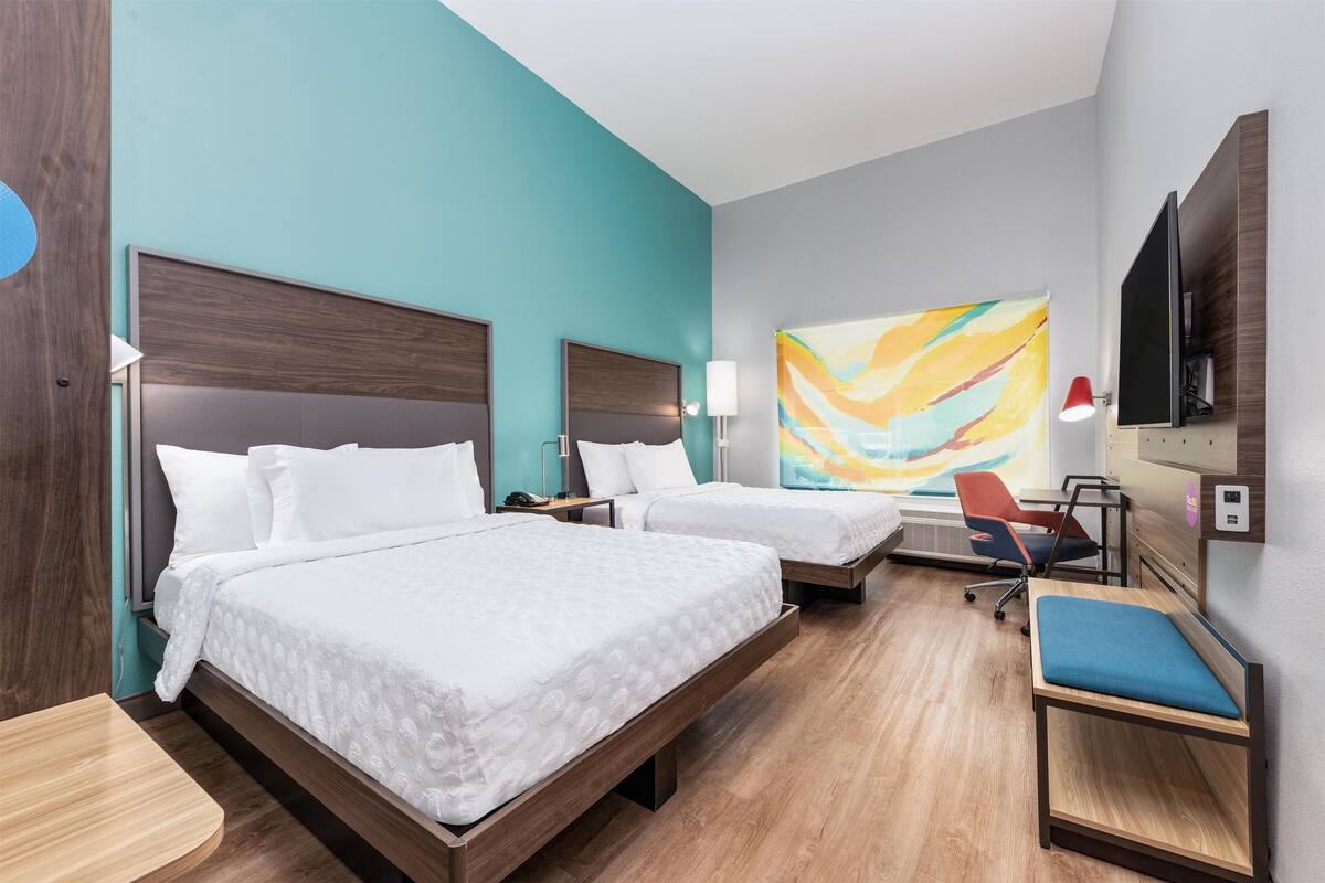 A hotel room with two beds, a desk, a chair, a TV, and a colorful abstract painting on the wall.