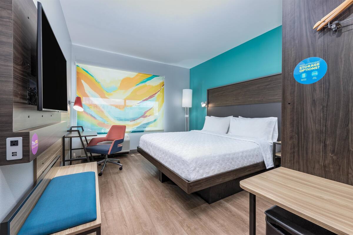 A modern hotel room with a bed, a desk, a chair, a TV, and a large abstract painting on the wall.