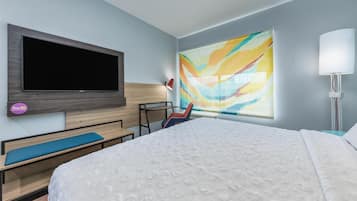 A hotel room with a large bed, a flat-screen TV mounted on the wall, a colorful abstract painting, a desk with a chair, and a bedside lamp.