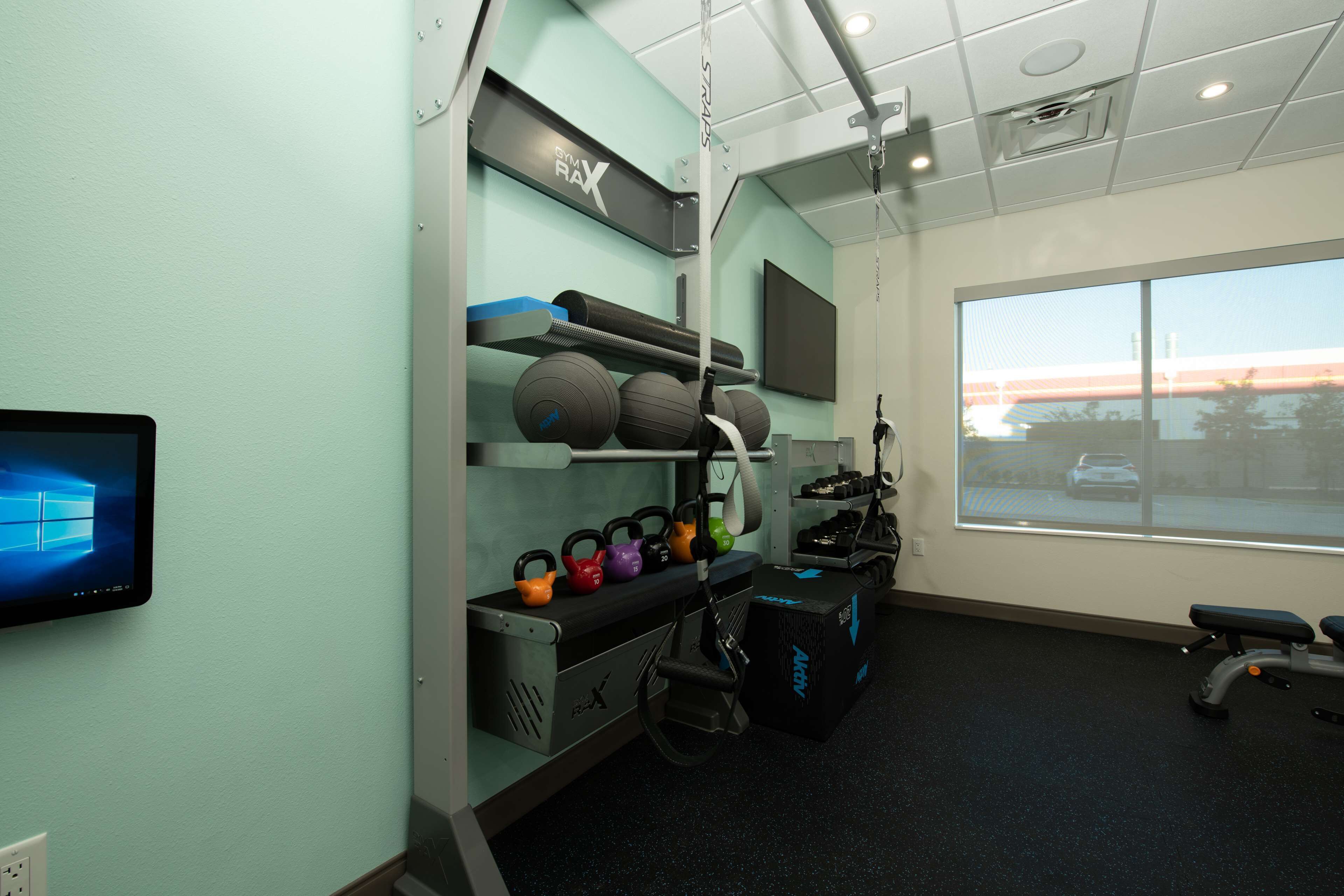 Ein Fitnessstudio mit Trainingsgeräten, einem an der Wand montierten Fernseher und einem Fenster mit Blick auf einen Parkplatz.