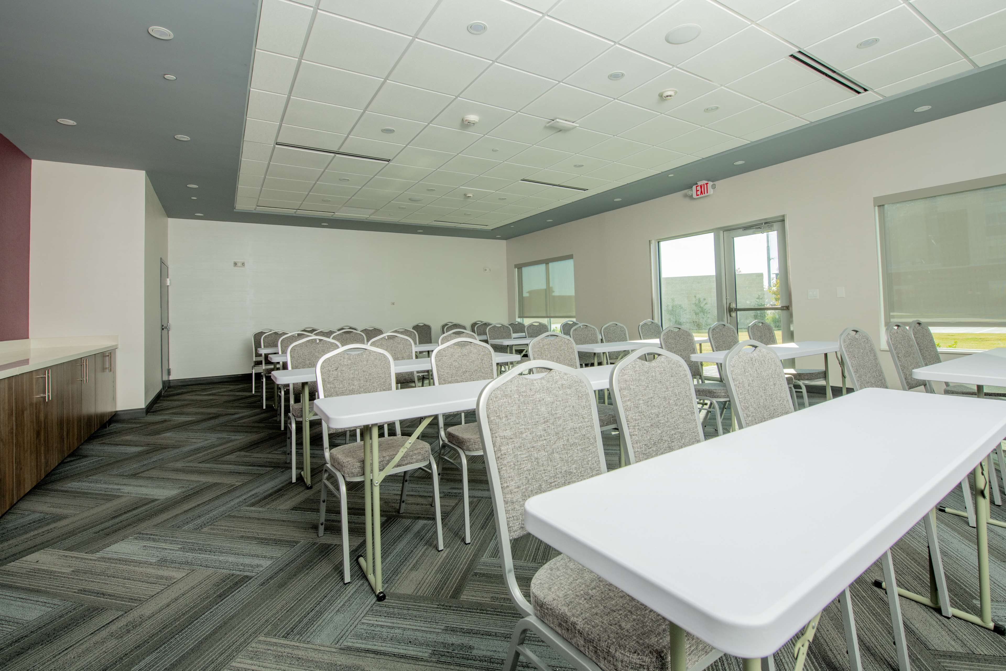 Une salle de conférence avec des tables et des chaises blanches, un sol recouvert d’un tapis gris et une fenêtre laissant entrer la lumière naturelle.