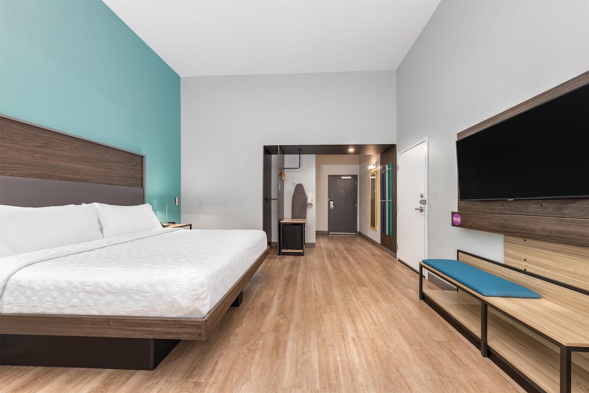 A modern hotel room with a large bed, a bench, a flat-screen TV, and a door leading to a hallway.