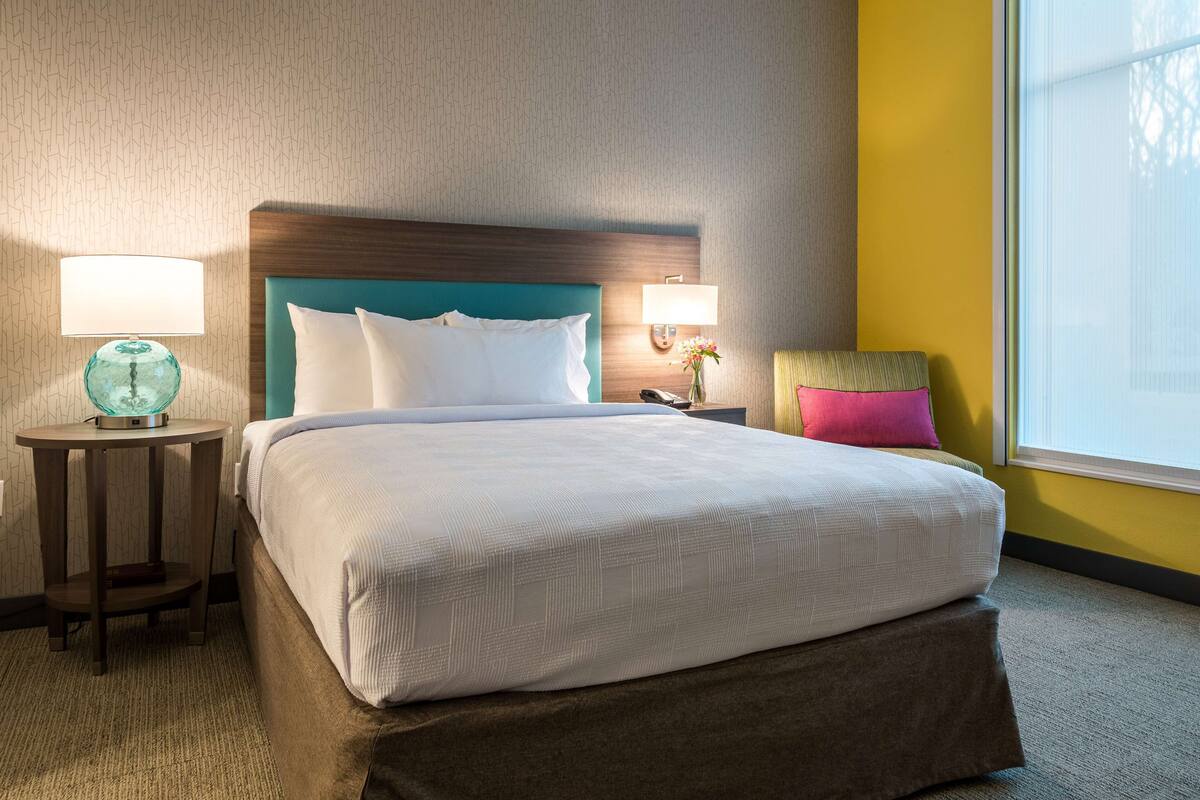 Une chambre d’hôtel avec un grand lit, deux tables de chevet, une petite table et une fenêtre avec des stores.